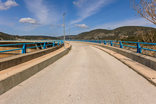 Road over a dam with blue railings and a mountainous landscape under a clear sky