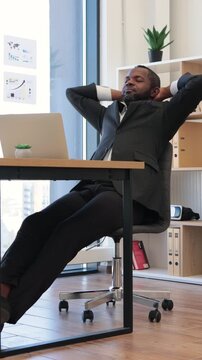 An African American man in a suit leans back in his office chair with his hands behind his head, looking content and successful