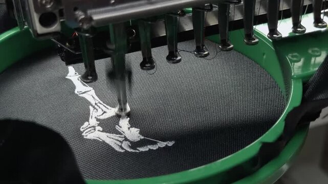 Extreme Macro Close Up of Professional Industrial Multi-Needle Embroidery Machine Head Stitching White Design on Black Fabric in Garment Factory Workshop Handheld Authentic Production Scene