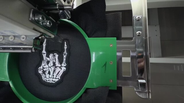 High Angle Top Down View Industrial Embroidery Machine Stitching White Skeleton Hand Rock Sign Design on Black Fabric Handheld Slow Motion Authentic Production