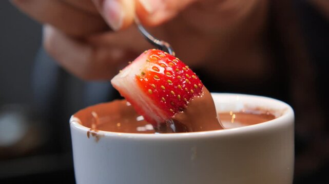 A hand using a small fork to dip a fresh strawberry into a white cup of melted chocolate.
