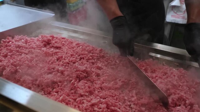 A close-up shot of a professional chef wearing black gloves using metal spatulas to stir-fry and brown a large amount of raw minced beef on a hot commercial stainless steel griddle.