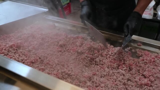 A close-up shot of a worker in black gloves using a large spatula to stir and mix a large quantity of raw ground beef in a stainless steel industrial container or mixer. Steam rises from the meat, sug