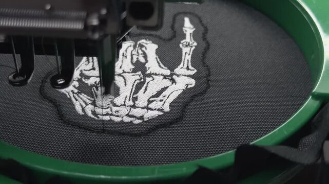 Macro Side View Industrial Computerized Embroidery Machine Stitching Final White Thread Border Outline on Skeleton Hand Rock Sign Design Black Fabric Handheld Slow Motion Authentic Production