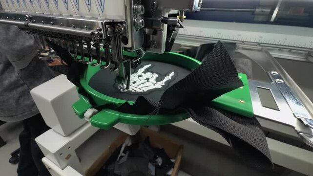 Wide Angle Perspective of Industrial Multi Needle Embroidery Machine Stitching White Skeleton Hand Design on Black Fabric in Textile Workshop Handheld Slow Motion Authentic Scene
