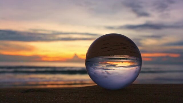 A glass sphere placed on the beach reflects an inverted view of a vibrant sunset over the ocean, creating a striking contrast between the warm golden sky and the deep blue clouds within the orb