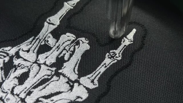 Macro Close Up Industrial Embroidery Machine Finishing White Skeleton Hand Rock Sign Design on Black Fabric Handheld Slow Motion Authentic Production