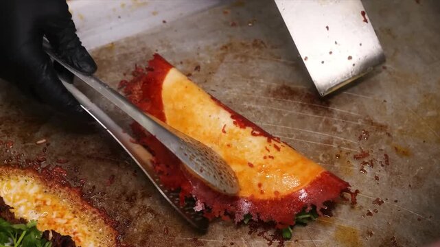 A professional chef, wearing black gloves, uses a metal spatula to fold a crispy, golden-brown cheese lace taco shell. The shell is filled with vibrant fresh cilantro and seasonings, being prepared on