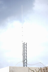 Obraz premium Tall, slender telecommunications antenna and metal lattice base mounted on a concrete roof against a cloudy sky. Modern broadcasting infrastructure and wireless communication concept.