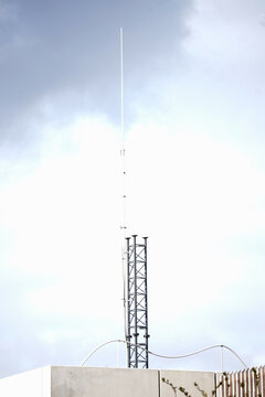 Tall, slender telecommunications antenna and metal lattice base mounted on a concrete roof against a cloudy sky. Modern broadcasting infrastructure and wireless communication concept.