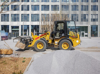 compact yellow wheel loader with a front bucket sits on a gravel surface during landscaping or site preparation work in front of a large modern office building 