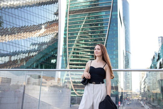 Young beautiful brunette woman wearing beige pants, black tank top, small bag staning outdoor on a city street near a tall glass office building. Trendy casual summer or spring outfit. Street fashion.