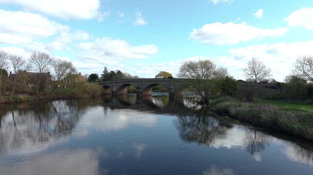 Wide landscape view of multi arch stone bridge with small car crossing over river with glassy reflections and village houses. Scenic rural roadway with historic structure and open sky. Bridge crossing