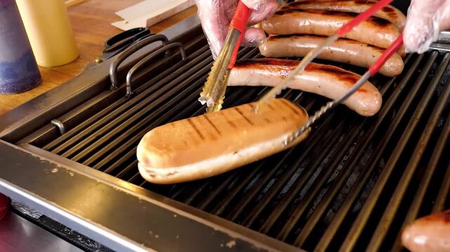 A close-up shot of several sausages and a single hot dog bun being grilled on a hot, professional-style barbecue grill. Tongs are visible in the background, and condiments are blurred in the foregroun