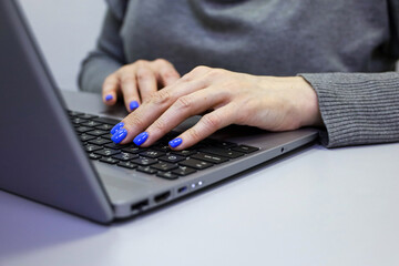 Woman in grey blouse working on the laptop keyboard sitting at table. Female hands with blue manicure