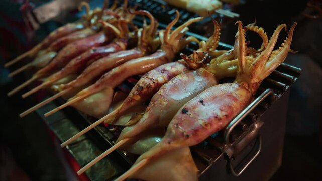 Close-up view of fresh whole squid on bamboo skewers being grilled over hot charcoal at a street food stall