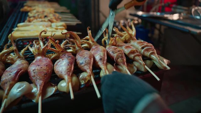 Close-up of a street food vendor using scissors to cut grilled squid on skewers at a night market in Bangkok, Thailand