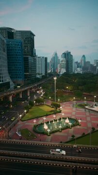 Vertical aerial view of Lumphini Park entrance with illuminated fountains, city traffic, and modern Bangkok skyline at dusk, Thailand
