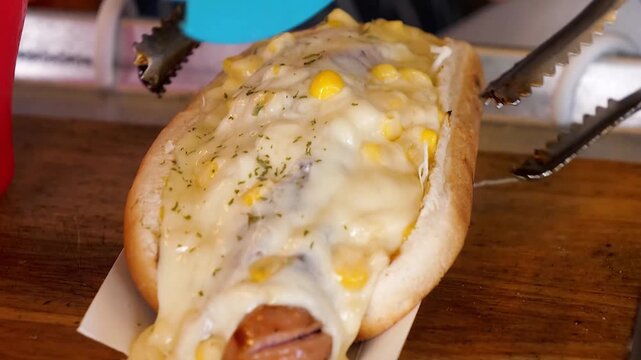 A close-up shot of a gourmet hot dog in a bun, generously topped with a melted cheese sauce and corn kernels. The fast food item is being held with tongs over a wooden surface, ready to be enjoyed.