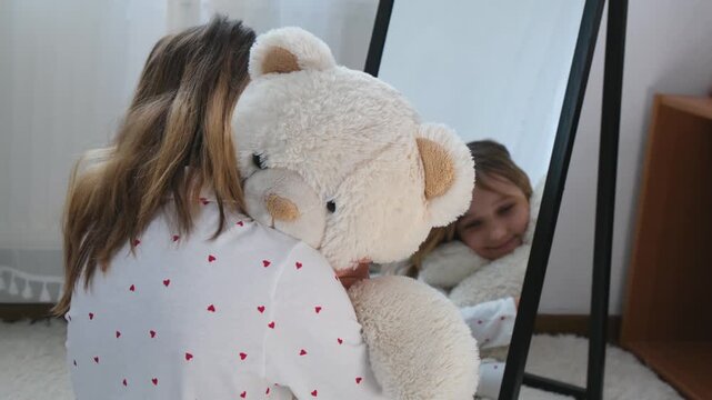 Happy little girl playing with teddy bear at home in front of mirror. Child enjoying imagination and role play, concept of joyful childhood, comfort and playtime.