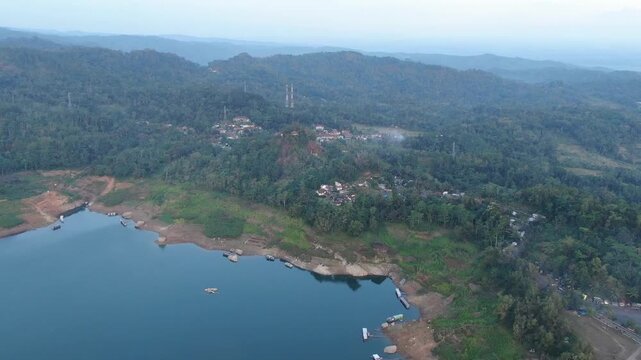 A sweeping high-angle drone shot captures the vast blue waters of Wadaslintang Reservoir surrounded by lush green hills