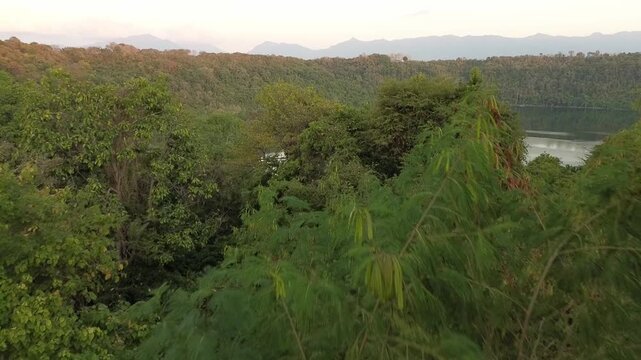 A cinematic drone reveal shot rising over a dirt path and thick vegetation to uncover the hidden Danau Asmara nestled within a dense green valley