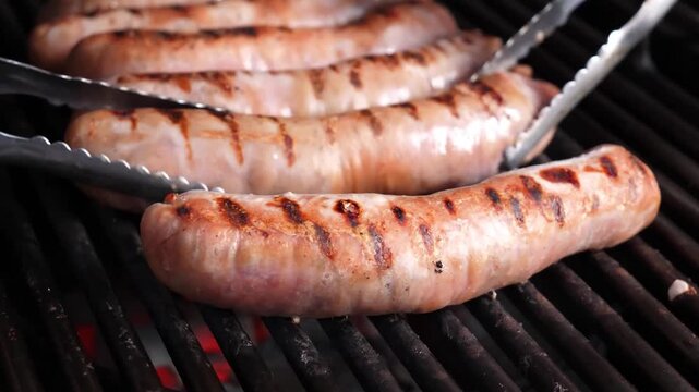 Close-up shot of juicy, raw sausages being grilled on a hot, black metal grate over an open flame. A hand uses metal tongs to turn the sausages, which are developing appealing brown grill marks. The s