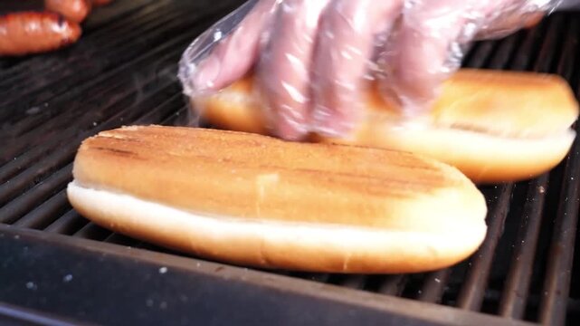 High-angle shot of a chef's hand in a plastic glove placing fresh hot dog buns onto a hot, steaming grill alongside rows of juicy, charred sausages.
