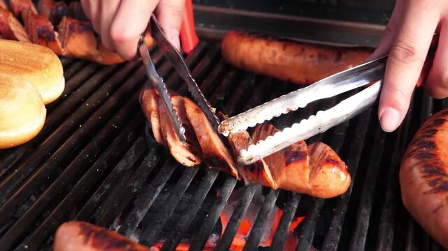 Close-up footage of sliced traditional Polish sausages, known as kielbasa, being grilled over intense heat on a cast iron barbecue grate. Tongs are used to turn the sizzling meat, highlighting the coo
