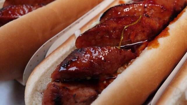 A close-up shot of a perfectly grilled, scored sausage (or hot dog) nestled in a soft bun, coated in a glossy barbecue or chili sauce. The image captures the appetizing texture of the meat and the sau