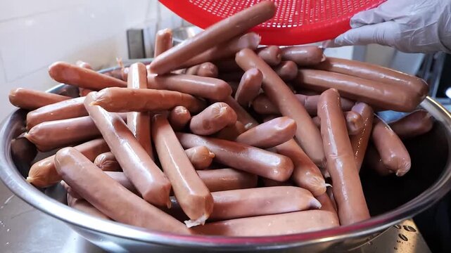A close-up shot of a kitchen worker wearing white gloves and a black uniform, holding a vibrant red plastic basket filled with a large quantity of fresh, raw frankfurter sausages.