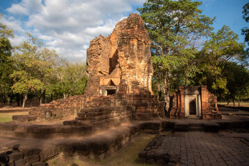Fototapeta premium Ancient brick ruins at Khao Khlang Nok in Thailand, a historic archaeological site surrounded by trees under a blue sky with clouds