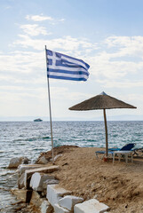 Sandy beach in Greece with straw umbrellas and lounge chairs near the calm sea, framed by a fluttering Greek flag