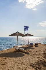 Sandy beach in Greece with straw umbrellas and lounge chairs near the calm sea, framed by a fluttering Greek flag