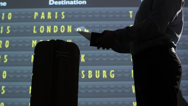 Traveler silhouette in airport lounge checks departure board for paris and london while holding passport, ticket and luggage, planning an international journey and upcoming flight