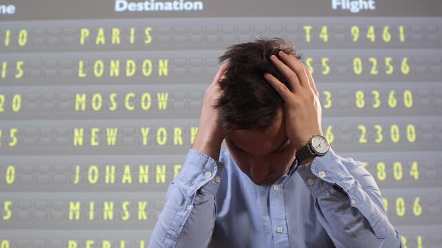 Stressed man covering his head with hands, reacting to a delayed or canceled flight on an airport display board, feeling anxiety and frustration about travel interruption