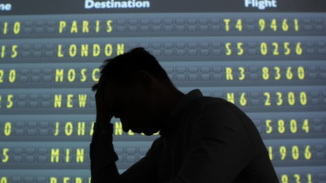 Stressed man silhouette sitting in an airport terminal with hands on his face, staring at a departure board of international flights travel delay, missed connection, exhaustion and frustration