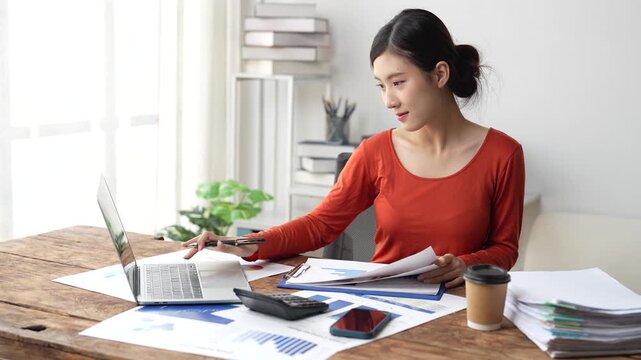 Young asian businesswoman sitting at her desk working on a laptop computer, checking financial reports and paperwork, preparing a project and smiling while working in office