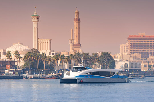 12 January 2026, Dubai, UAE: Modern RTA ferry boat traveling across Creek with traditional minarets, palm trees, and historic architecture lining the shore