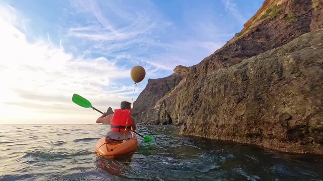 Kayaking ocean cliffs. A man in a life vest paddles an orange kayak with a balloon along a rocky coast under clear skies.