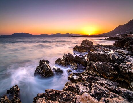 A picturesque seaside scene at sunset. The rocky shore contrasts with the calm waves and vibrant colors of the evening sky