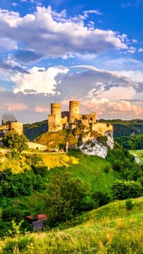 A picturesque stone castle ruins sits atop a grassy hill under a partly cloudy sky. The landscape is lush and vibrant