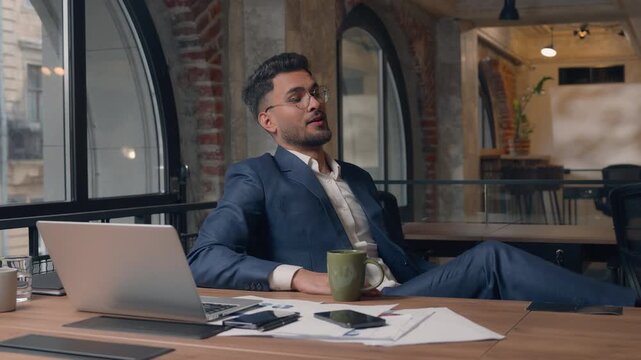 Arabian Indian business man laptop job done businessman resting drinking coffee putting legs on office table successful employer guy male end task finishing chatting online complete computer browsing