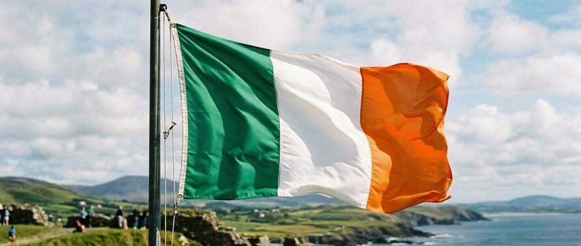 Irish tricolor flag waving from a flagpole above rugged coastal cliffs, overlooking the Atlantic Ocean and rolling green fields under a cloudy sky &mdash; a windswept national symbol.