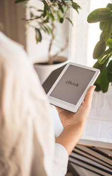 Woman holding ebook reader home near books and plants in warm sunlight. Digital reading, cozy lifestyle, self education, modern technology in comfortable home atmosphere, mindful slow living moments