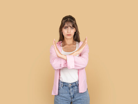 Serious caucasian woman making stop gesture with crossed arms, expressing rejection or refusal concept idea image, wearing pink shirt, isolated on beige studio background with copy space.