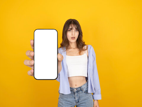 Surprised caucasian woman holding smartphone with blank screen mockup toward camera, presenting device concept, wearing striped shirt and denim, isolated on yellow studio background with copy space.