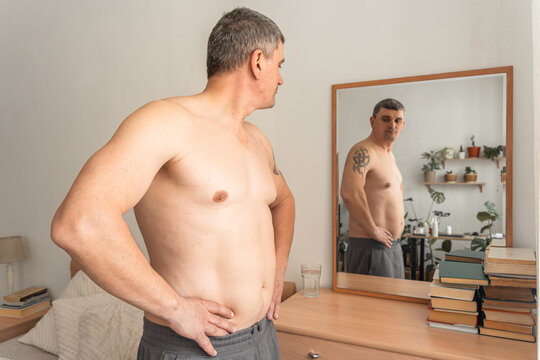Mature man standing shirtless, looking at his reflection in a mirror, examining his changing body shape and waistline. Real person, self-acceptance, authentic lifestyle