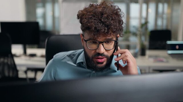 Arabian business man looking at computer monitor talking mobile phone at office ethnic businessman focused look in pc screen talk smartphone discussing online work result male using internet at office