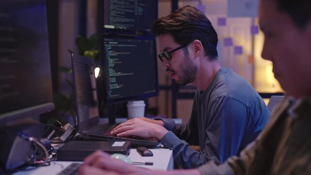 Male software engineer concentrating on his computer screens while working overtime in a dark room.
